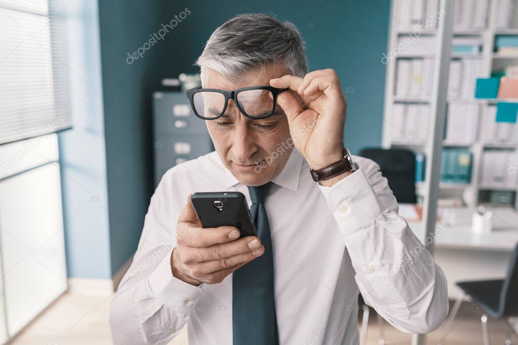 Corporate businessman using his smartphone and having vision problems, he is adjusting his glasses