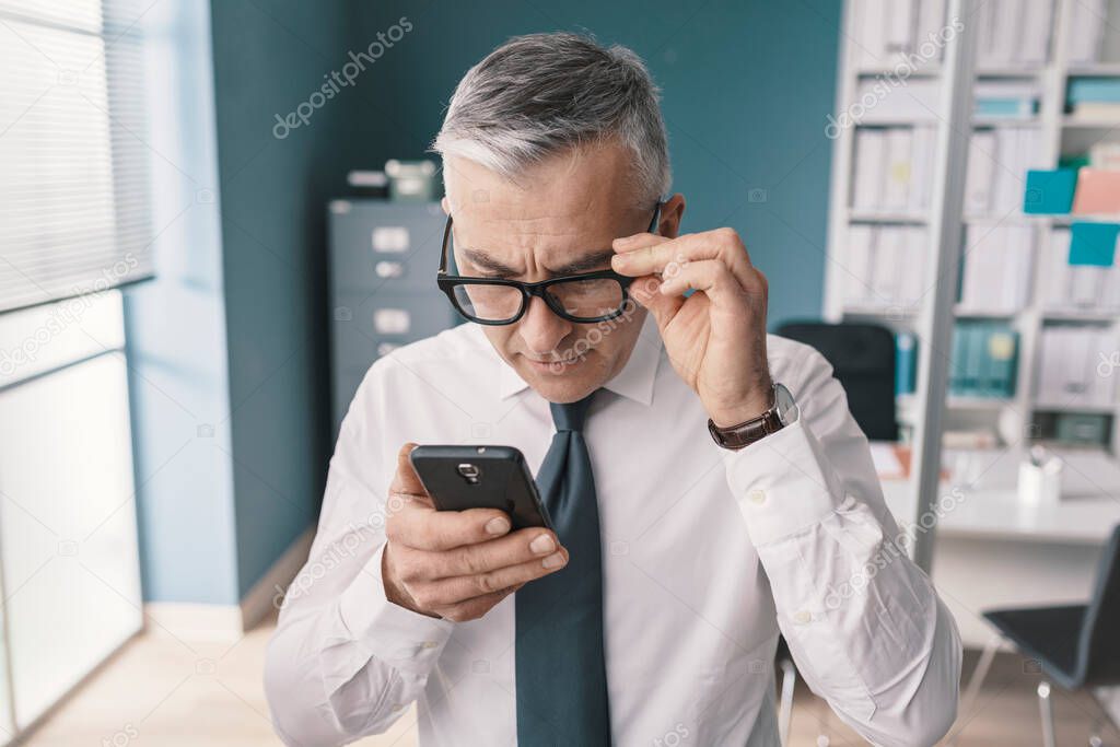 Corporate businessman using his smartphone and having vision problems, he is adjusting his glasses
