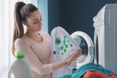 Çamaşır makinesinin yanında duran ve çamaşır yıkadıktan sonra leke yapmış elbiseleri tutan hayal kırıklığına uğramış ev hanımı.
