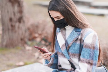 Yüz maskeli genç bir kadın parkta oturmuş akıllı telefonuyla sohbet ediyor.