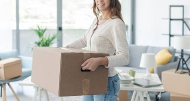 Yeni evine taşınan mutlu genç kadın, yanında bir karton kutu taşıyor.