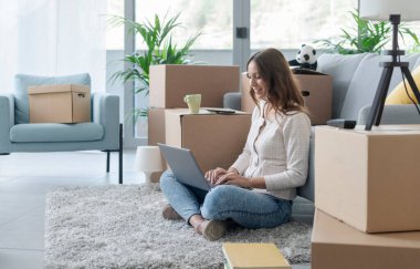 Yeni dairesine taşınan mutlu genç kadın yerde oturuyor ve laptopuyla bağlantı kuruyor.