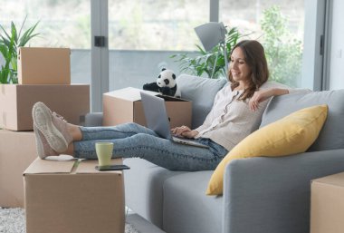 Yeni dairesine taşınan mutlu genç kadın koltukta oturuyor ve laptopuyla bağlantı kuruyor.