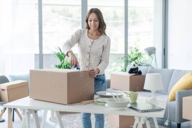 Mutlu genç bir kadın yeni evine taşınıyor. Bir karton kutuyu kesiciyle açıyor.