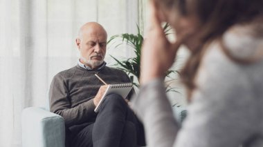 Psikoterapi seansı sırasında hastasını dinlerken notlar yazan profesyonel bir terapist.