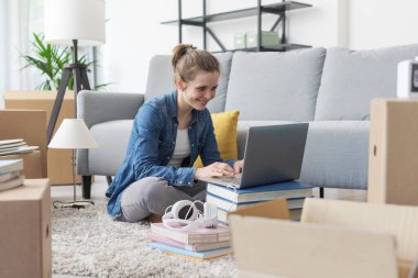 Neşeli genç bayan yeni dairesine taşınıyor, yerde oturuyor ve laptopuyla bağlantı kuruyor.