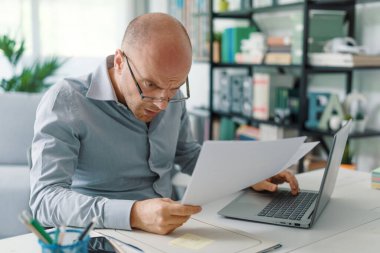 Hayal kırıklığına uğramış işadamı masasında oturup evrakları kontrol ediyor, evden çalışıyor.