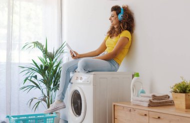 Çamaşır makinesinde oturup çamaşırların bitmesini bekleyen mutlu genç kadın tabletine bağlanıyor ve kulaklık takıyor.