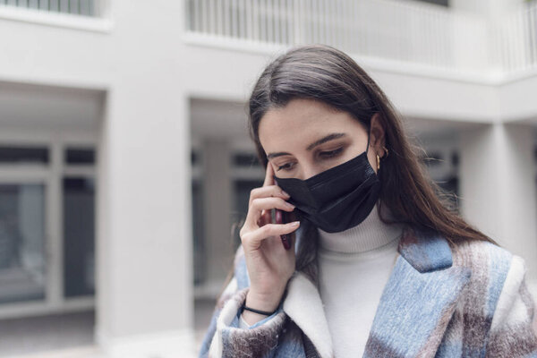 Young woman walking down the street and having a phone call with her smartphone, she is wearing a face mask
