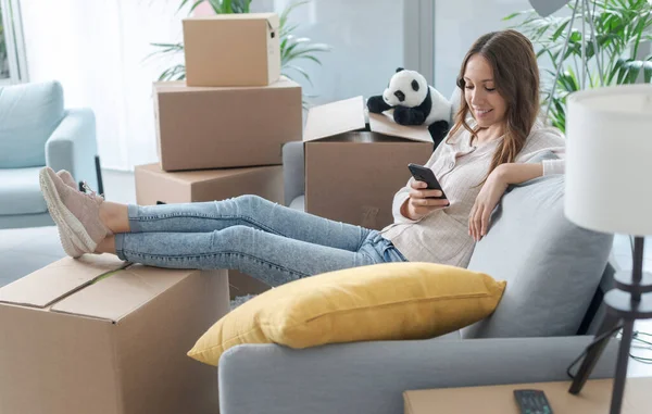 新しい家に引っ越してきた女性はソファに座ってスマートフォンと接続しています — ストック写真