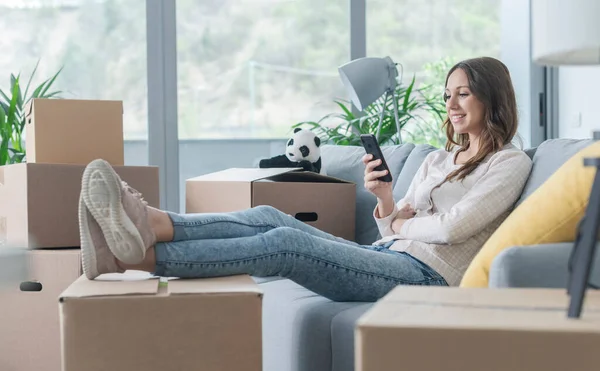 新しい家に引っ越してきた女性はソファに座ってスマートフォンと接続しています — ストック写真