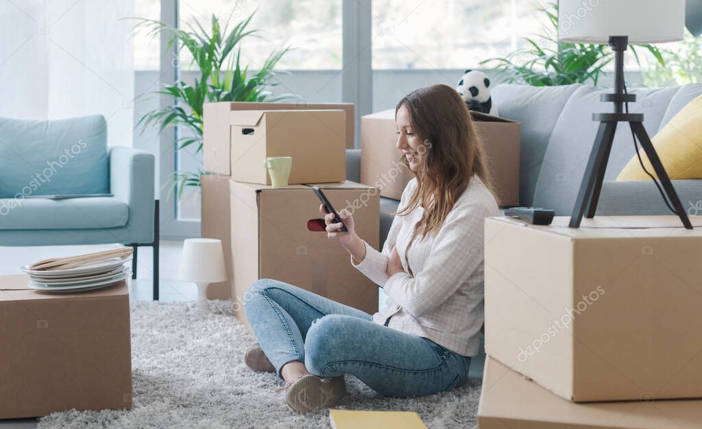 Happy woman in her new apartment, she is sitting on the floor and chatting with her smartphone