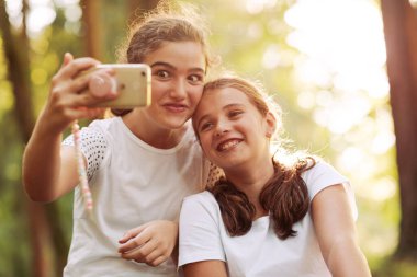 Ormanda birlikte poz veren tatlı genç kızlar akıllı telefonlarıyla selfie çekiyorlar.