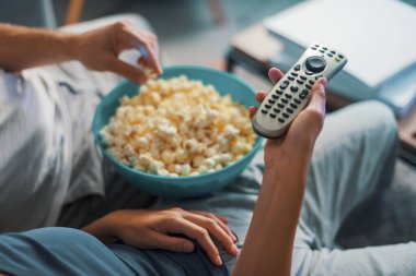 Çift kanepede oturup televizyon izliyor, kadın uzaktan kumandayı tutuyor ve adam patlamış mısır yiyor.