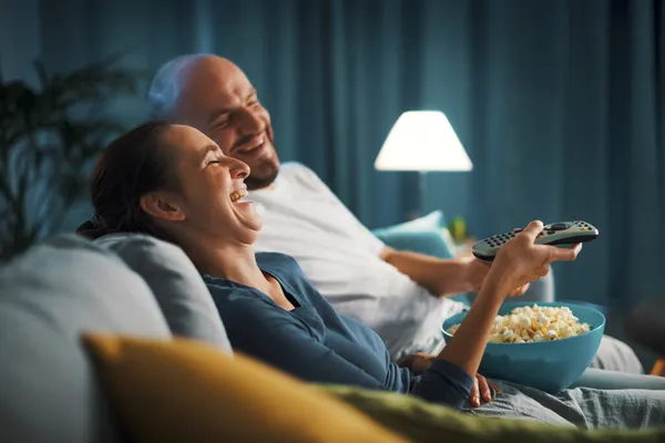 幸せなカップル座っているソファに自宅で 彼らは面白いコメディ映画を見ているとポップコーンを食べて — ストック写真