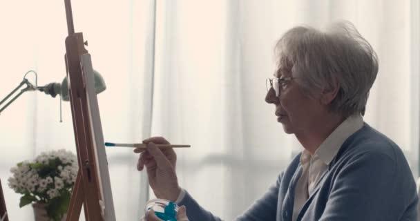 Creatieve senior vrouw zit aan het bureau en schildert op doek — Stockvideo