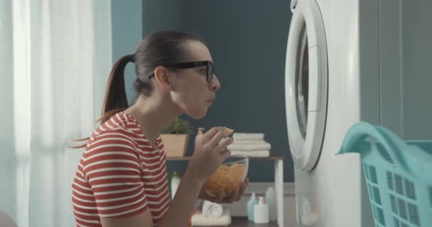 Vrouw die chips eet terwijl ze wacht tot de wasmachine klaar is — Stockvideo