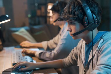 Teenager friends playing online video games together, they are wearing headsets and staring at the computer monitor