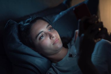 Young woman lying in her bed at night and connecting with her smartphone, she can't sleep