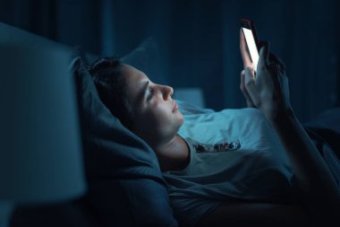 Young woman lying in her bed at night and connecting with her smartphone, she can't sleep
