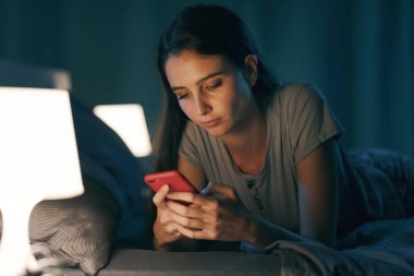 Sleepless young woman lying in bed at night and checking her smartphone
