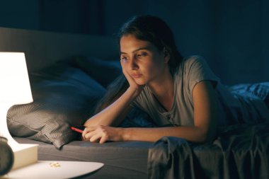 Sad young woman lying in bed and suffering from insomnia, she is holding her smartphone