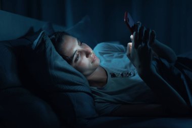 Young woman lying in her bed at night and connecting with her smartphone, she can't sleep