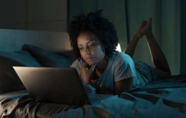 Young african american woman relaxing in bed at night, she is connecting online with her laptop