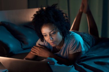 Young african american woman relaxing in bed at night, she is connecting online with her laptop