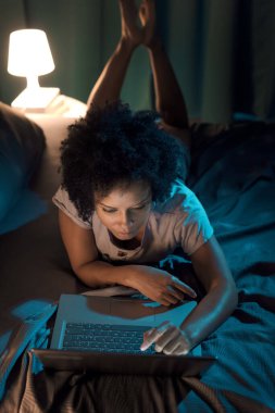 Young african american woman relaxing in bed at night, she is connecting online with her laptop