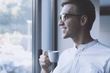 Man standing next to a window and looking away, he is having a coffee break