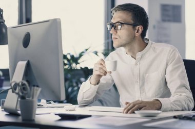 Businessman sitting at desk and working with his computer, he is drinking a coffee