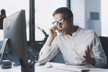 Angry nervous office worker sitting at desk and shouting on the phone