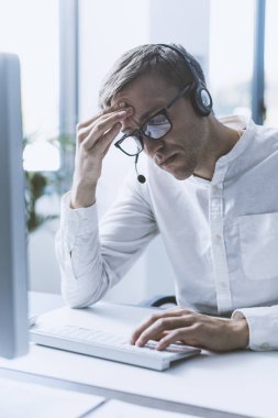 Exhausted office worker sitting at desk, he is tired and having a headache, job burnout concept