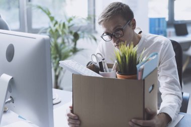 Fired office worker packing his belongings in a box and leaving the office, he is sitting at desk and thinking