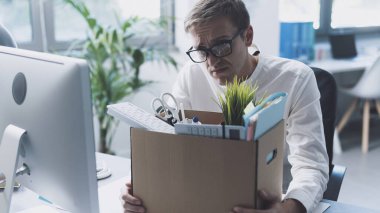 Fired office worker packing his belongings in a box and leaving the office, he is sitting at desk and thinking
