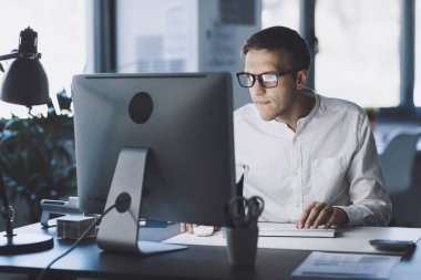 Young corporate businessman sitting at desk and working with his computer, he is focused and staring at the computer screen