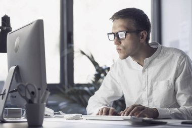 Young corporate businessman sitting at desk and working with his computer, he is focused and staring at the computer screen
