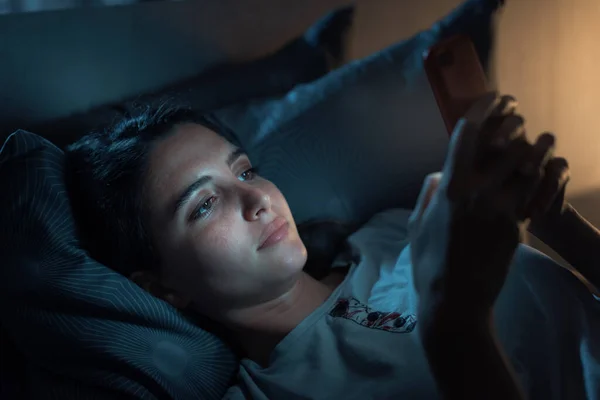 Young woman lying in her bed at night and connecting with her smartphone, she can\'t sleep