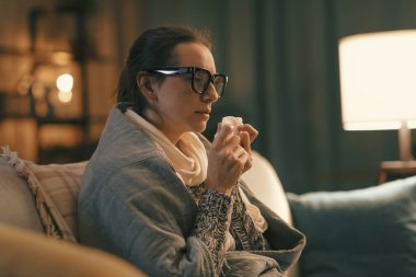 Sick woman with covid symptoms resting at home on the couch, she is blowing her nose