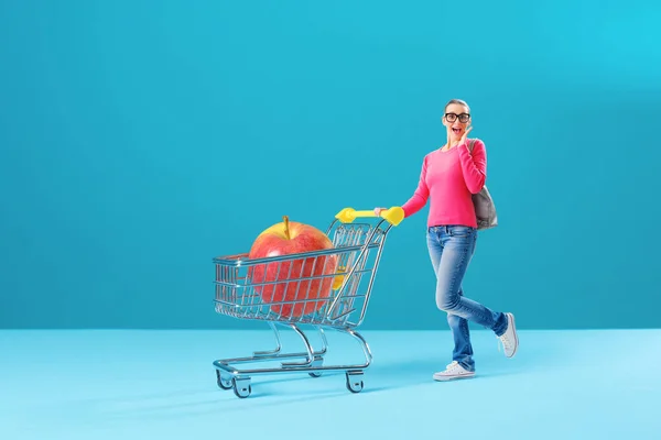 Tiny happy woman pushing a miniature shopping cart, she is buying an apple