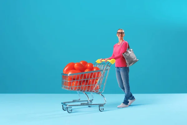 Tiny happy woman pushing a miniature shopping cart full of tomatoes