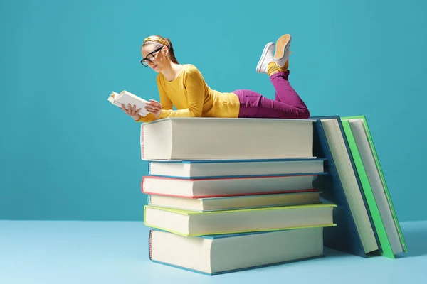 Tiny smart woman lying on a pile of books and reading, education and hobby concept