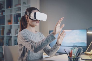 Young woman sitting at desk and wearing a VR headset, she is interacting with virtual reality