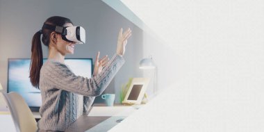 Young woman sitting at desk and wearing a VR headset, she is interacting with virtual reality, banner with copy space