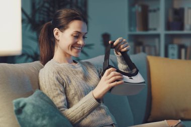 Smiling young woman sitting on the couch at home and wearing a VR headset, entertainment and virtual reality concept