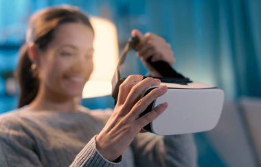 Smiling young woman sitting on the couch at home and wearing a VR headset, entertainment and virtual reality concept