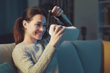 Smiling young woman sitting on the couch at home and wearing a VR headset, entertainment and virtual reality concept