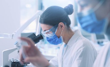 Young researcher looking at the samples under the microscope, she is wearing a face mask and protective equipment