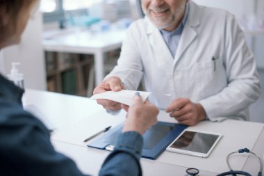 Smiling doctor sitting at desk and giving a medical prescription to his patient